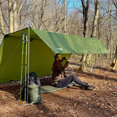 Man relaxing under DD Tarp 3x3 UV50+ in a forest, enjoying shade and comfort while sipping from a mug.